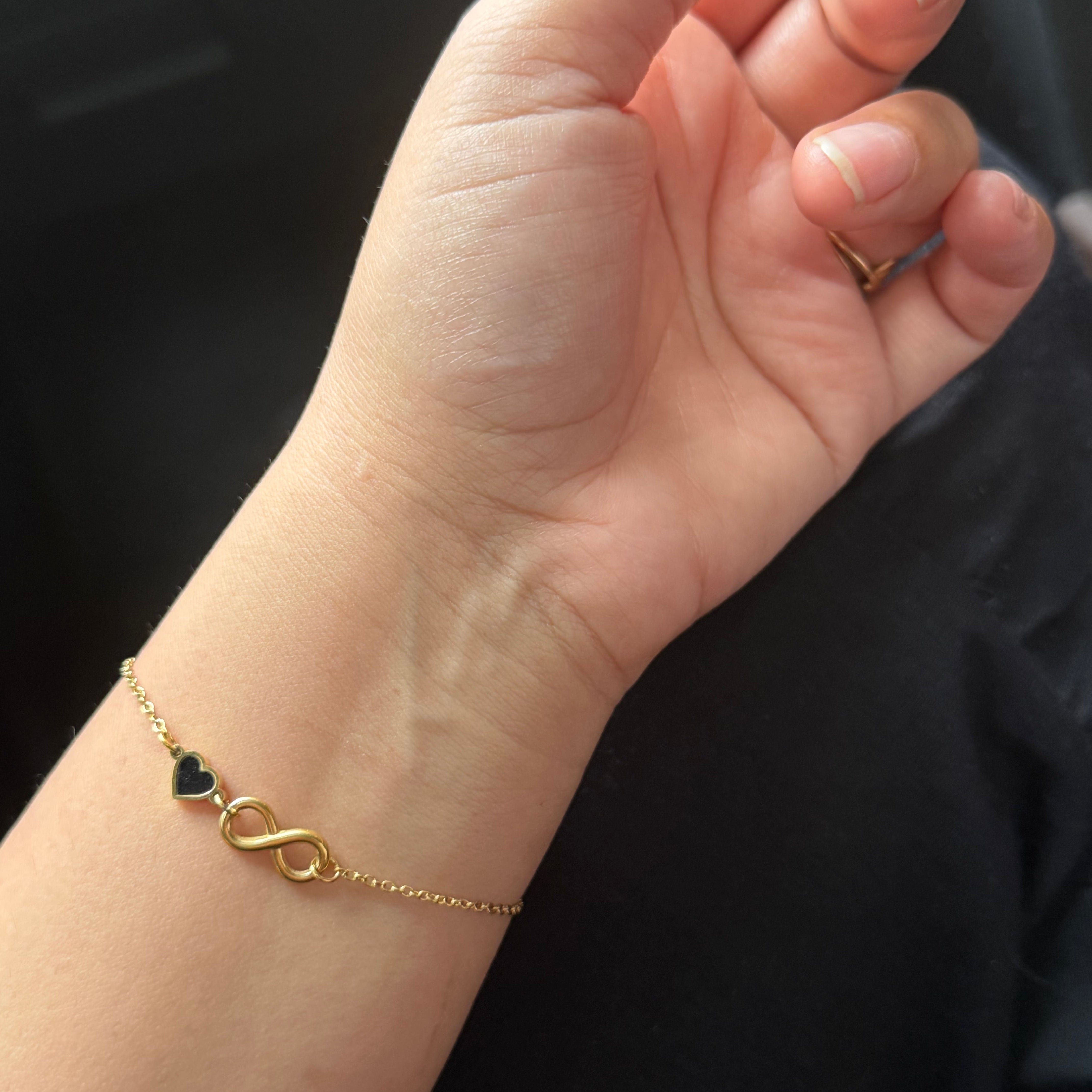 Gold bracelet with a heart and infinity symbol on a wrist against a dark background