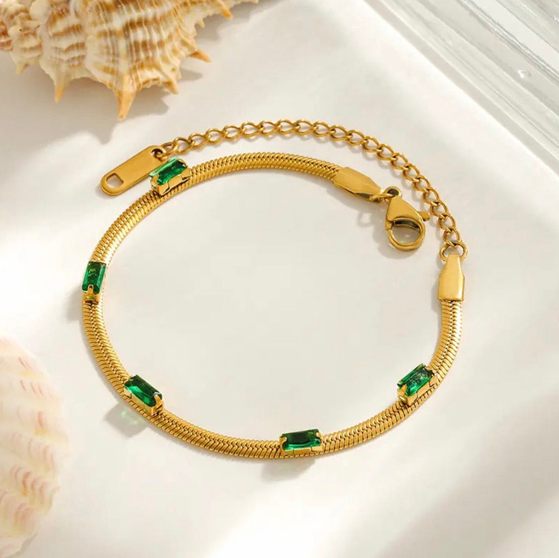Gold bracelet with green gemstones on a white surface with seashells.