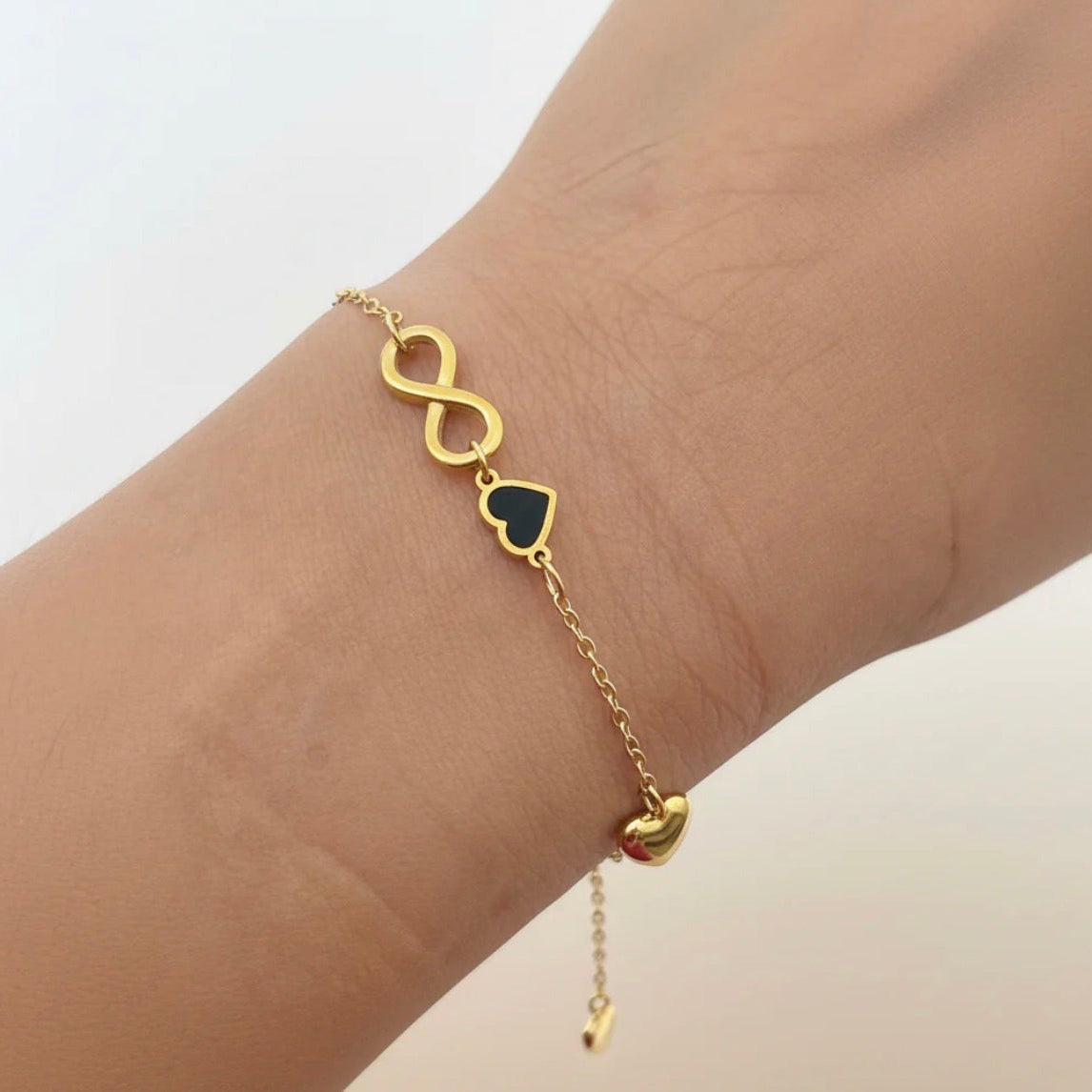 Gold bracelet with heart and infinity symbol on a wrist against a light background